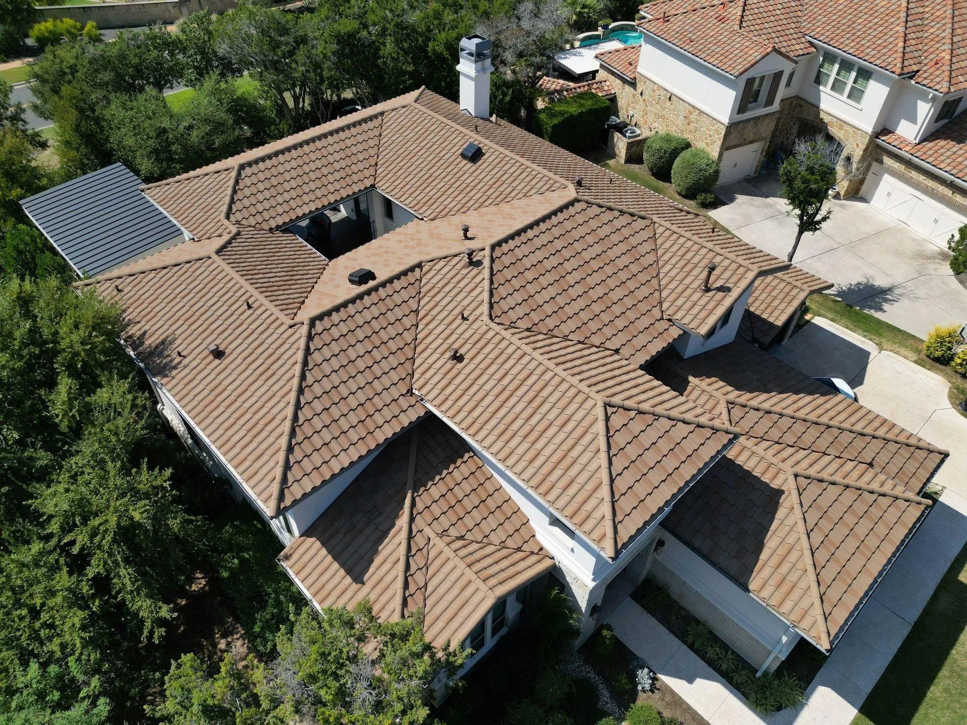 Aerial view of a large residential home with a complex, brown tiled hip roof surrounded by trees and a paved driveway.