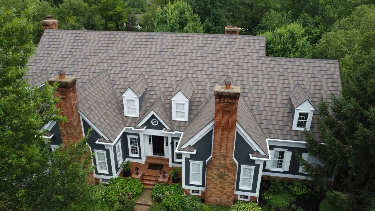 An aerial view of a multi-gabled house with grey siding, brick chimneys, white trim, and a brown shingled roof.