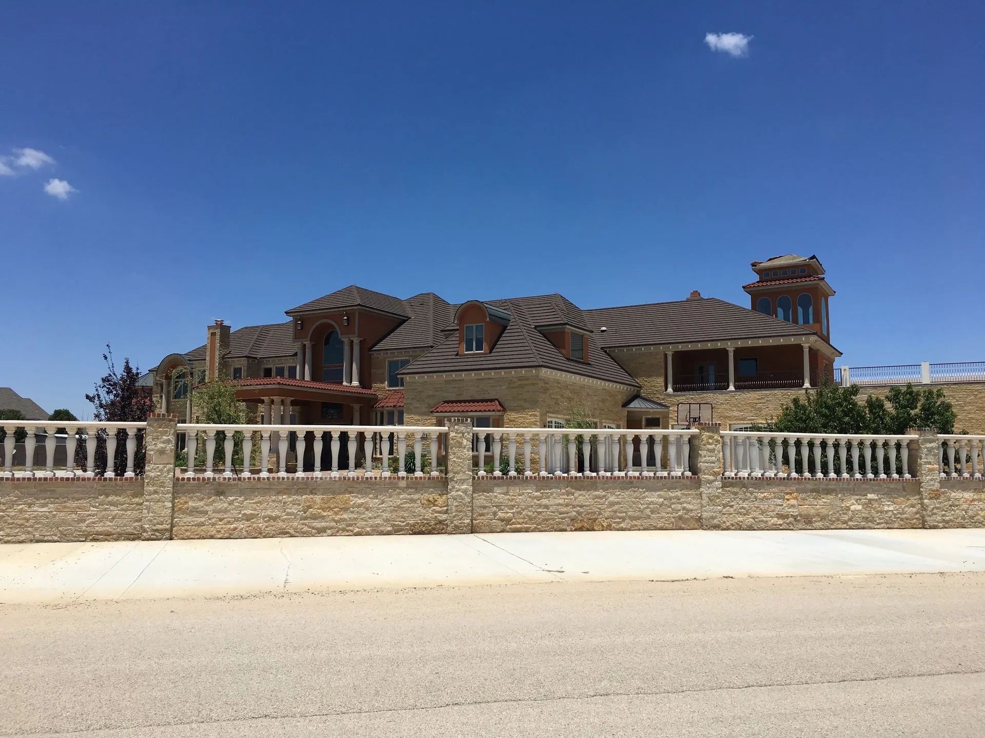 A large, two-story stone house with a complex roof and tower, enclosed by a stone and white balustrade fence under a blue sky.