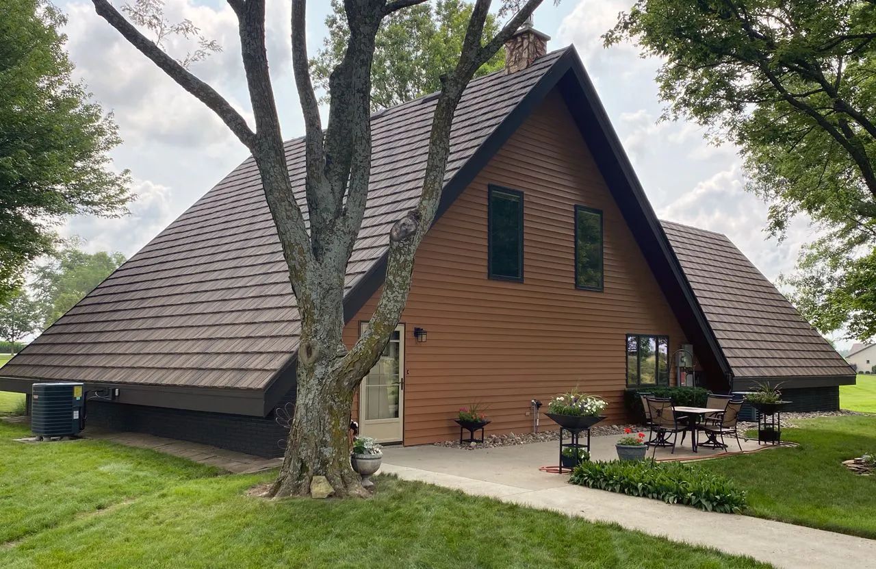 A brown A-frame house with a shingled roof, a patio with chairs, and a large tree in the front yard on a grassy lawn.
