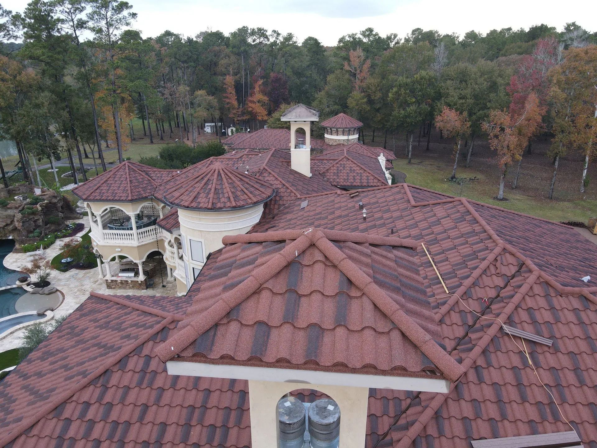Aerial view of a Mediterranean-style home with a dark red tiled roof, white stucco walls, and surrounding woodland.