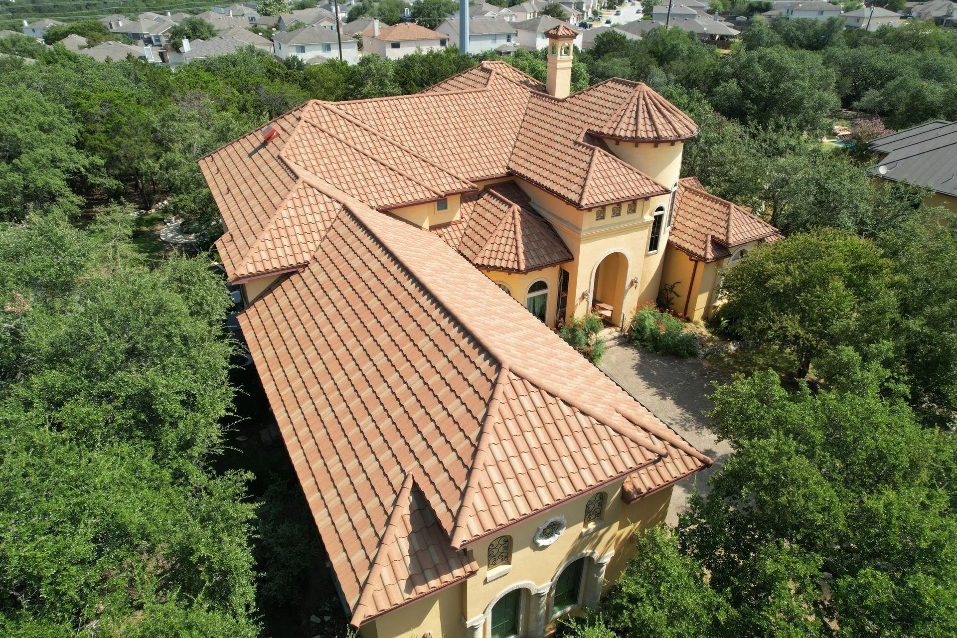 Aerial view of a large, tan-colored stucco home with a terracotta tiled roof, surrounded by dense green trees.