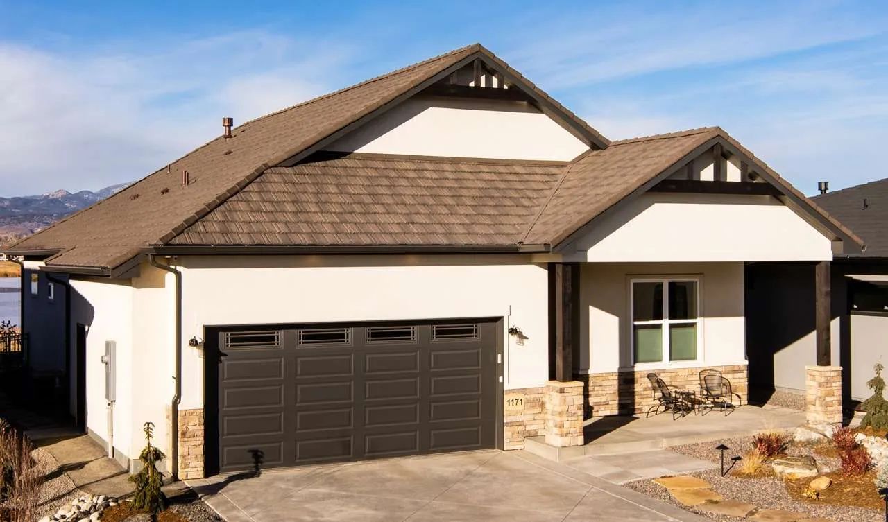 A single-story suburban house with a brown shingled roof, cream-colored stucco siding, stone accents, and a dark garage.