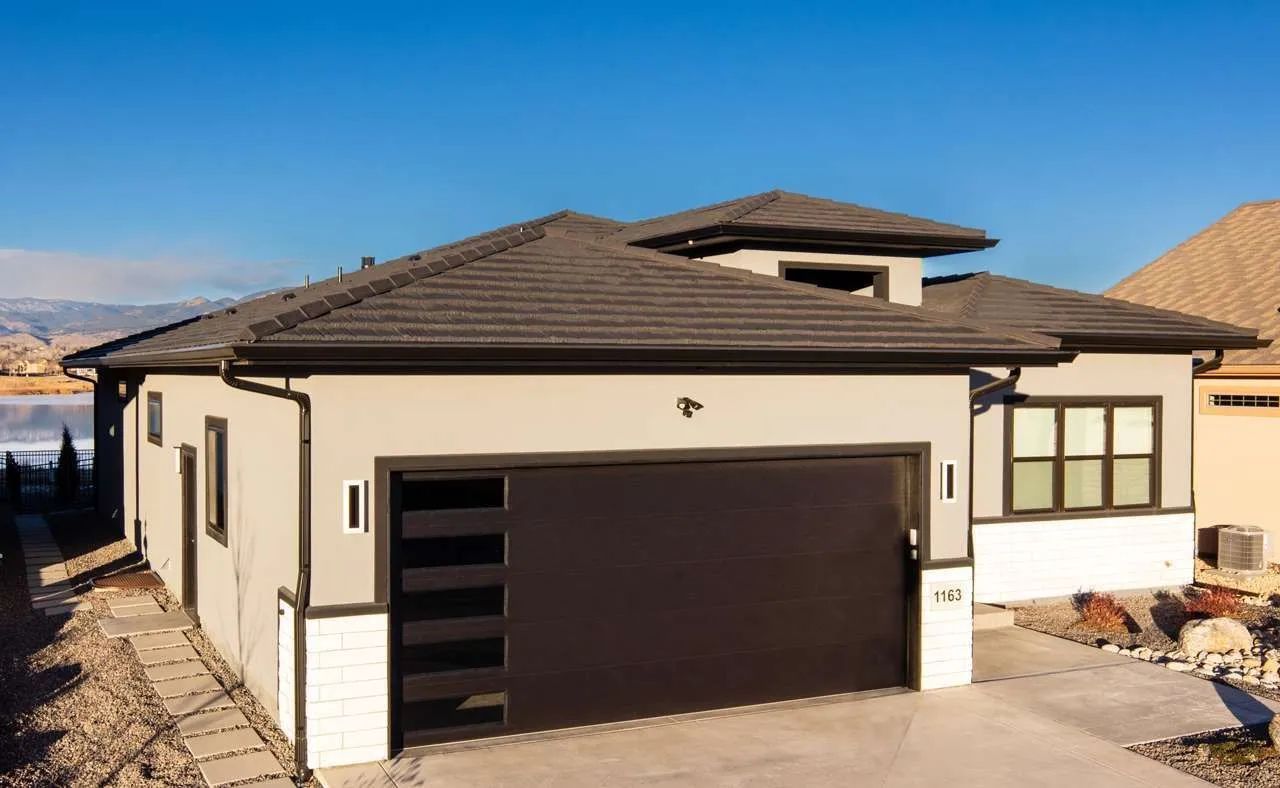 Single-story house with a grey stucco exterior, white brick accents, dark brown garage door, and a brown tiled roof.