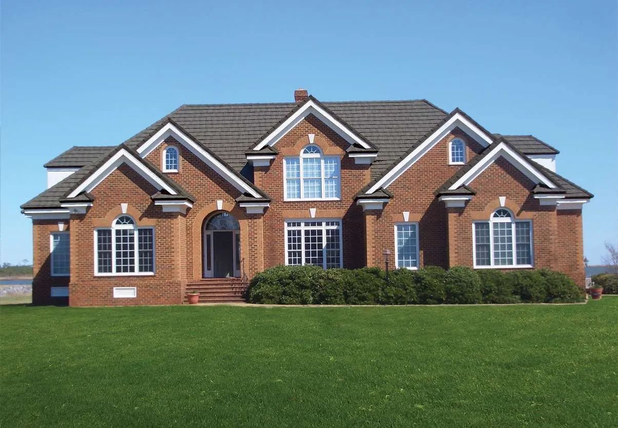 A two-story brick house with a dark gabled roof and a spacious green lawn under a clear blue sky.