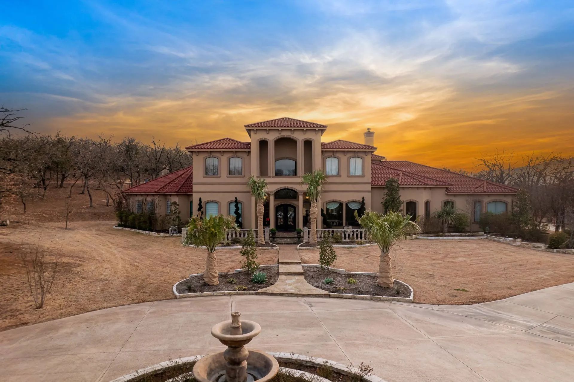 A two-story tan Mediterranean-style villa with a red tile roof stands behind a stone fountain at sunset.