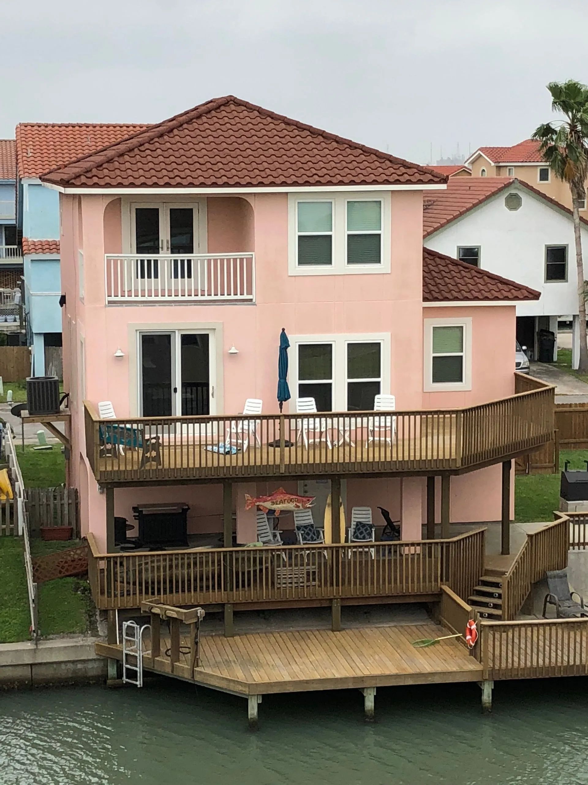 A multi-story pastel pink waterfront house with multiple wooden decks and a dock extending over the canal.