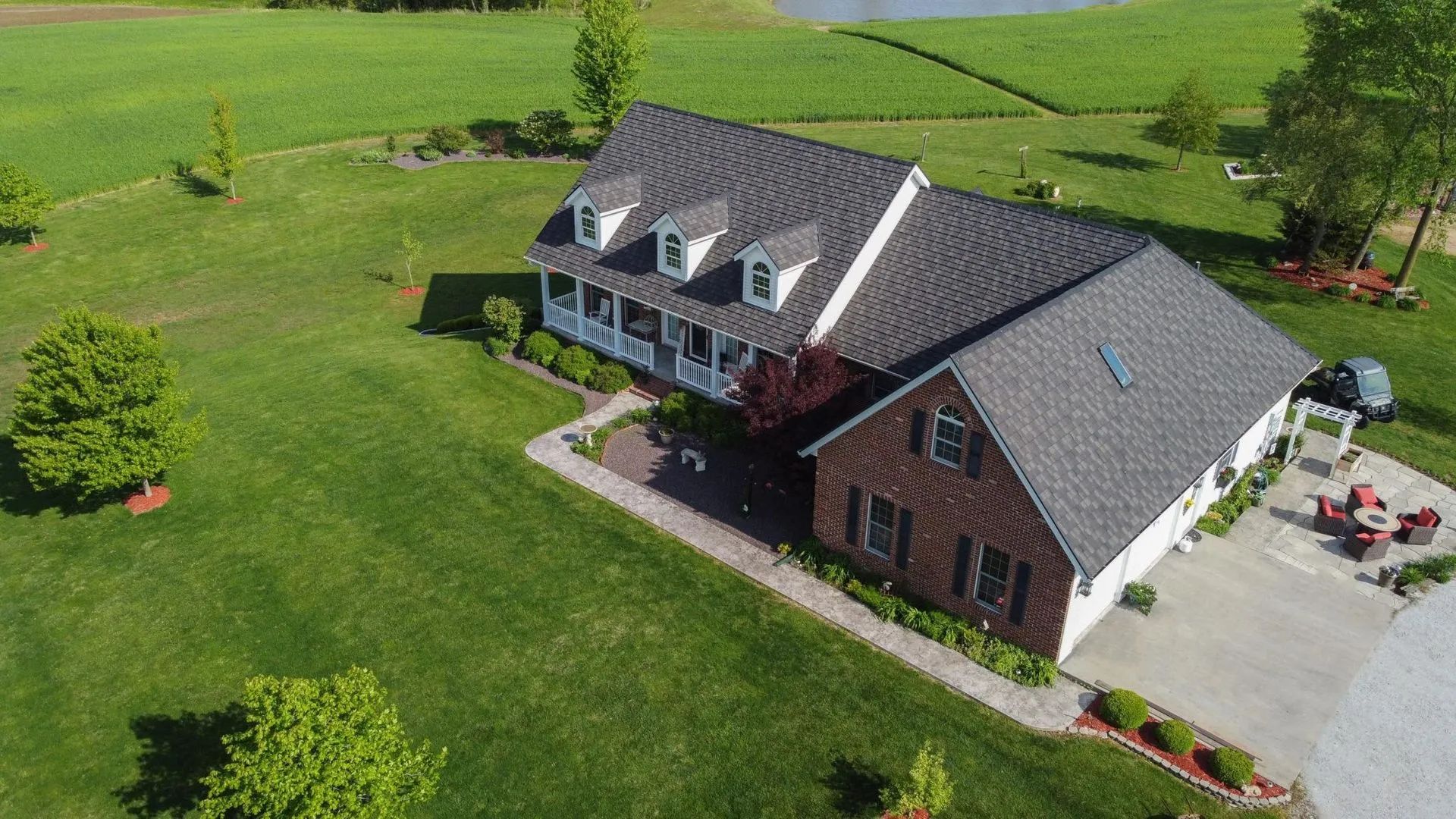 An aerial view of a brick house with a dark roof and dormer windows, surrounded by green lawns and patio space.