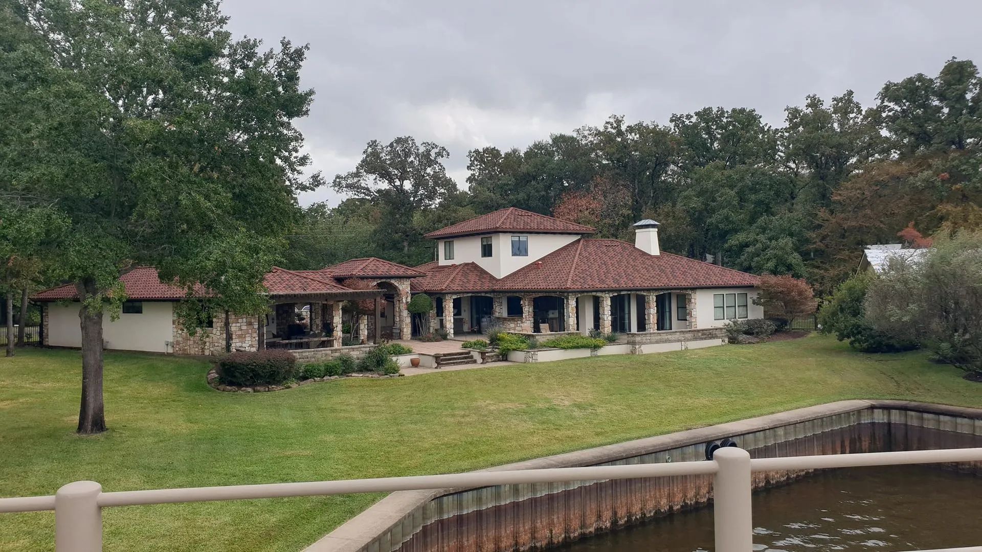 A white, Mediterranean-style house with a brown tile roof sits on a grassy lawn beside a lake with a wooden bulkhead.