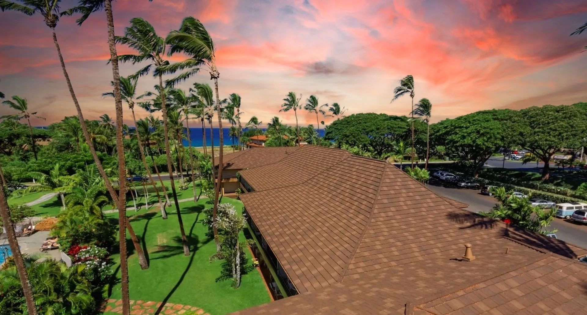 A high-angle view of a brown-roofed building surrounded by palm trees and lush grass at sunset overlooking the ocean.