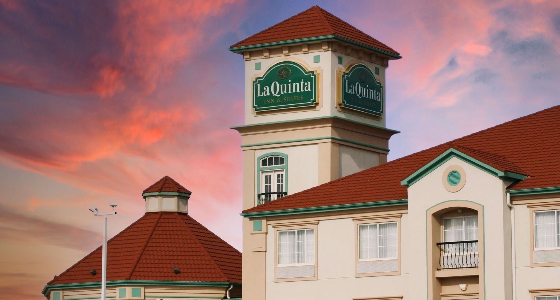 A La Quinta hotel building with a central tower and red tile roof, set against a pink and orange sunset sky.