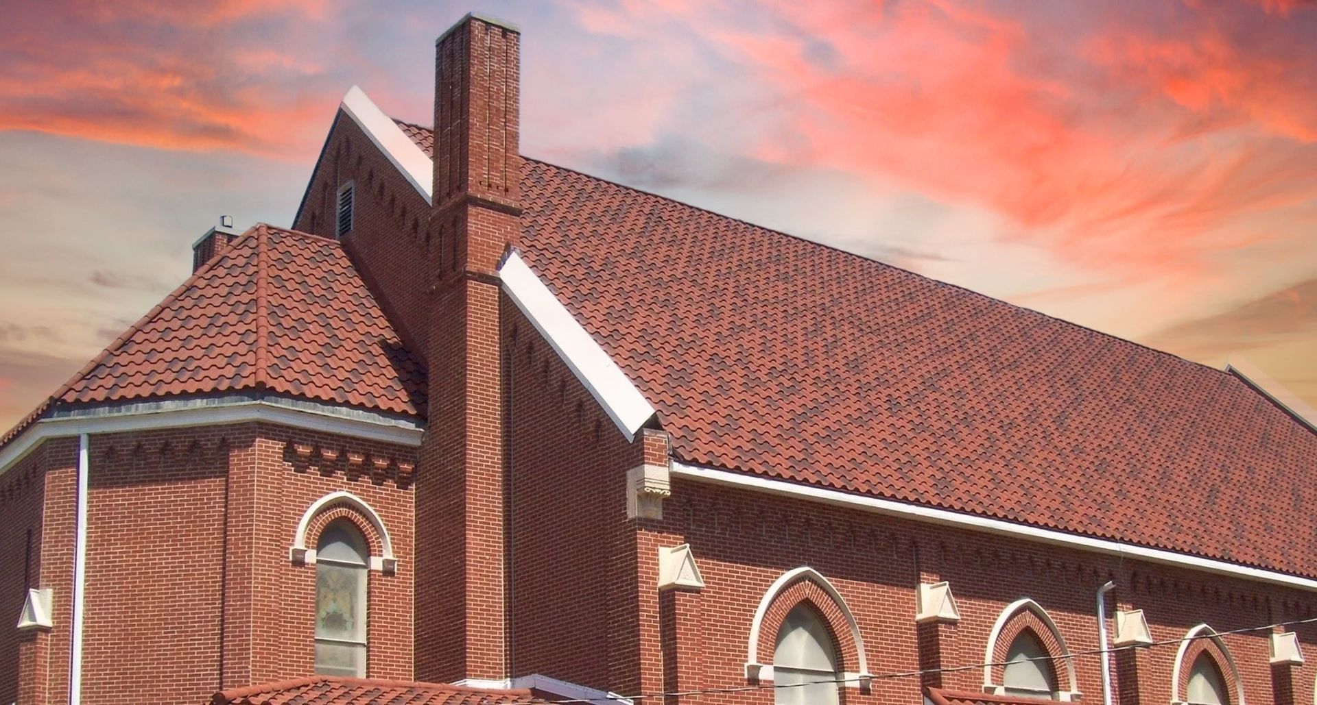 A red brick church building with arched windows and a roof against a sunset sky.