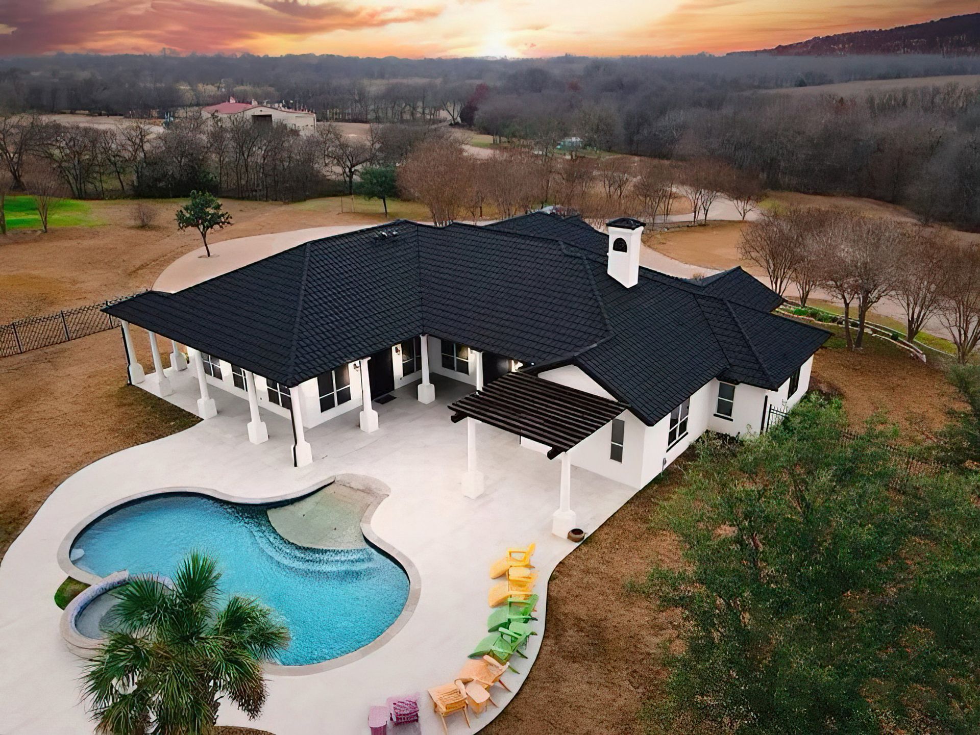 Aerial view of a white home with a black roof, swimming pool, and patio at sunset in a rural landscape.