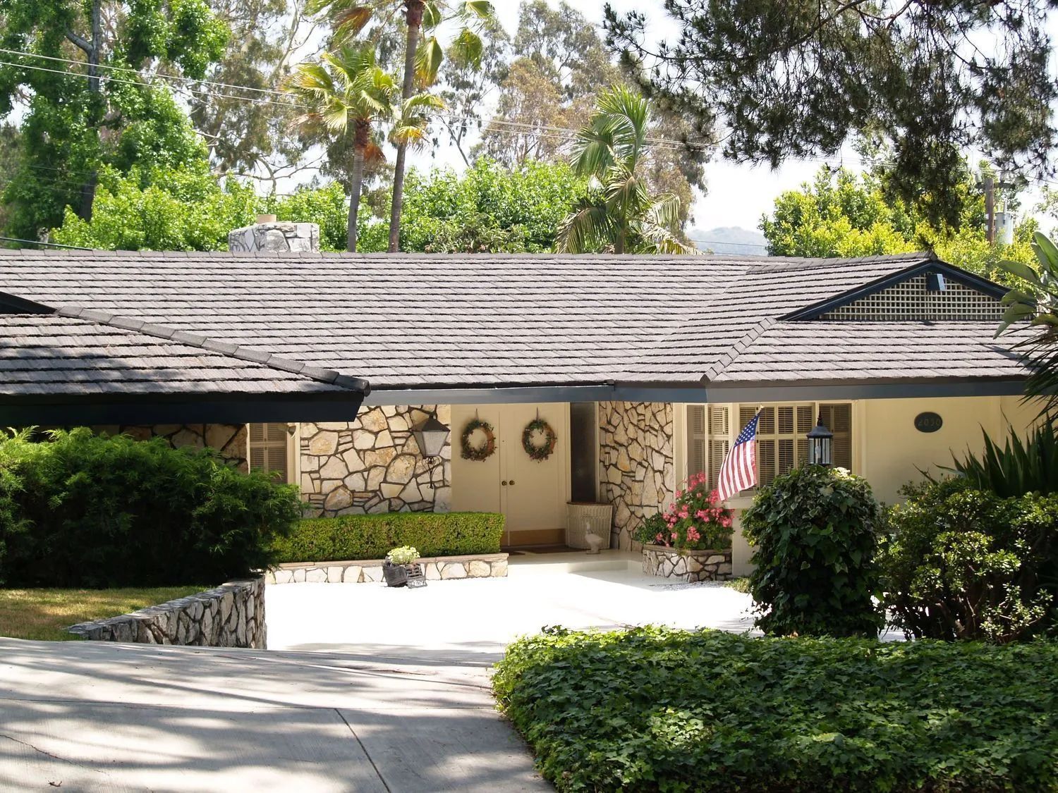 A low-slung, cream-colored house with stone accents and a dark-tiled roof, surrounded by trees and lush greenery.
