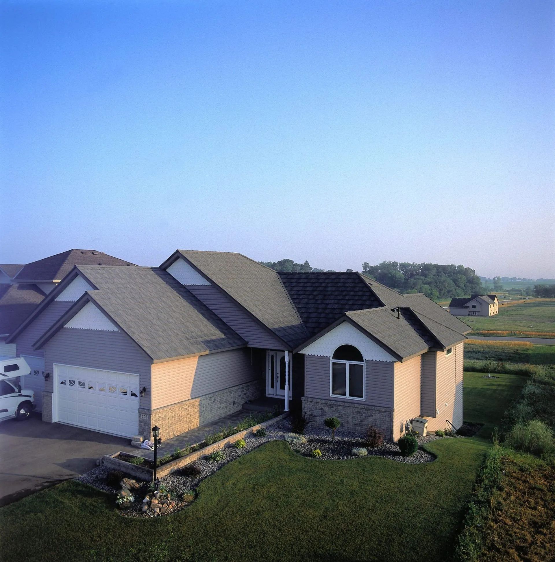 An aerial view of a single-story suburban house with a two-car garage, gray siding, stone accents, and a grassy lawn.