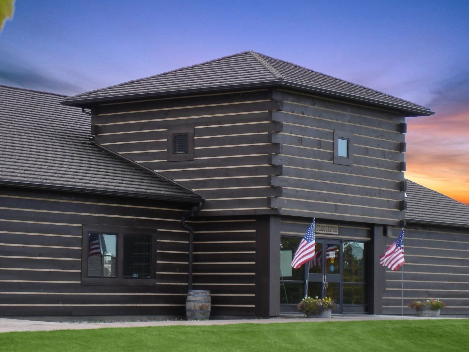 A dark brown, multi-level log building with American flags flanking the front entrance under a sunset sky.