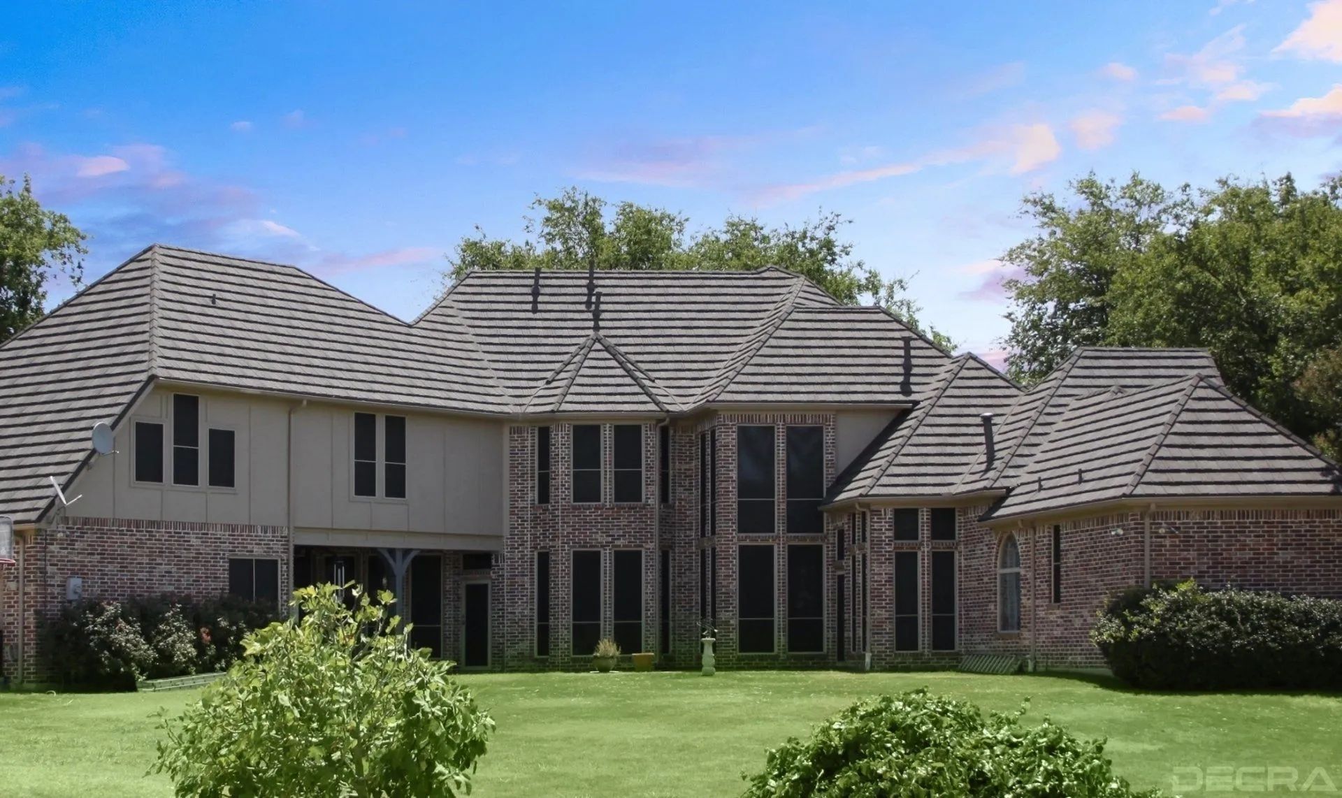 A large, two-story house with a grey tile roof and brick and tan siding, set behind a green lawn under a blue sky.