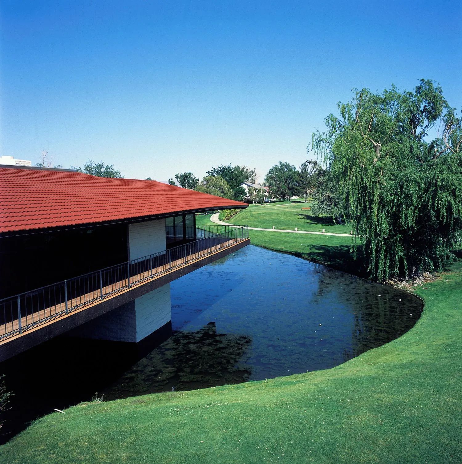 A modern building with a red tiled roof overlooks a pond and a grassy landscape under a clear blue sky.