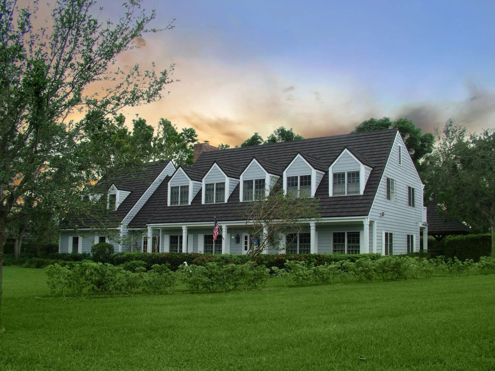A two-story white house with a dark roof and multiple gabled dormers stands behind a green lawn at sunset.