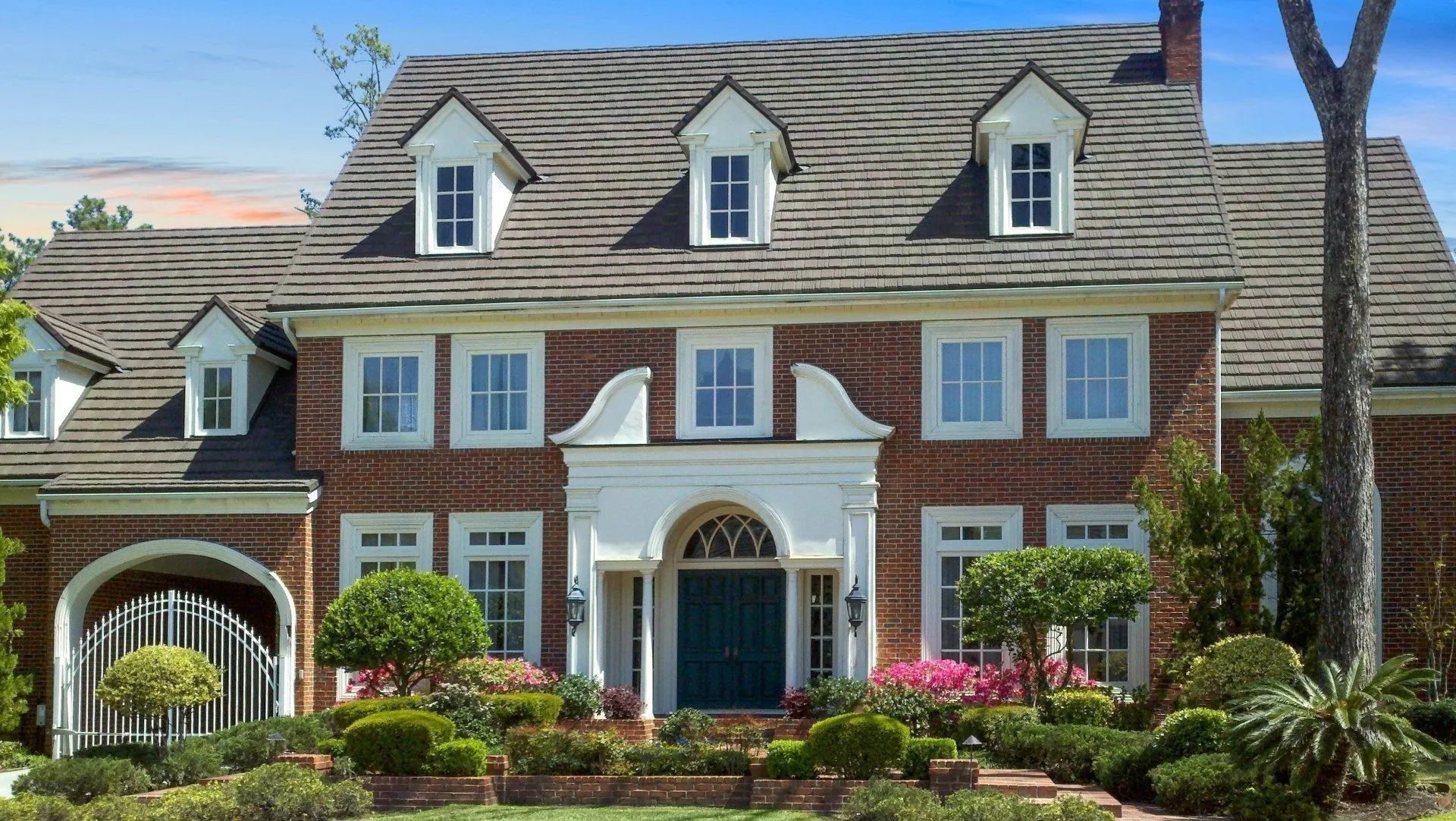 A two-story brick house with a dark blue front door, white arched entryway, dormer windows, and landscaped front garden.