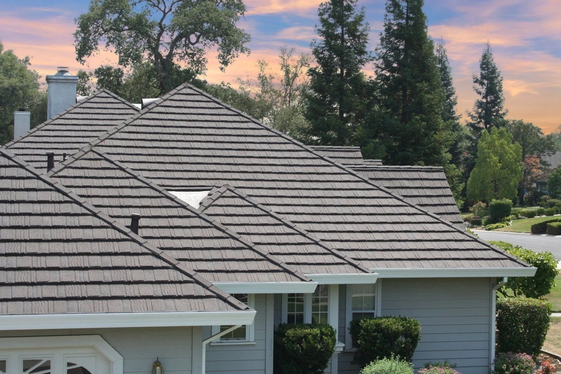 A multi-gabled house with a dark textured shingle roof under a sunset sky with surrounding trees.