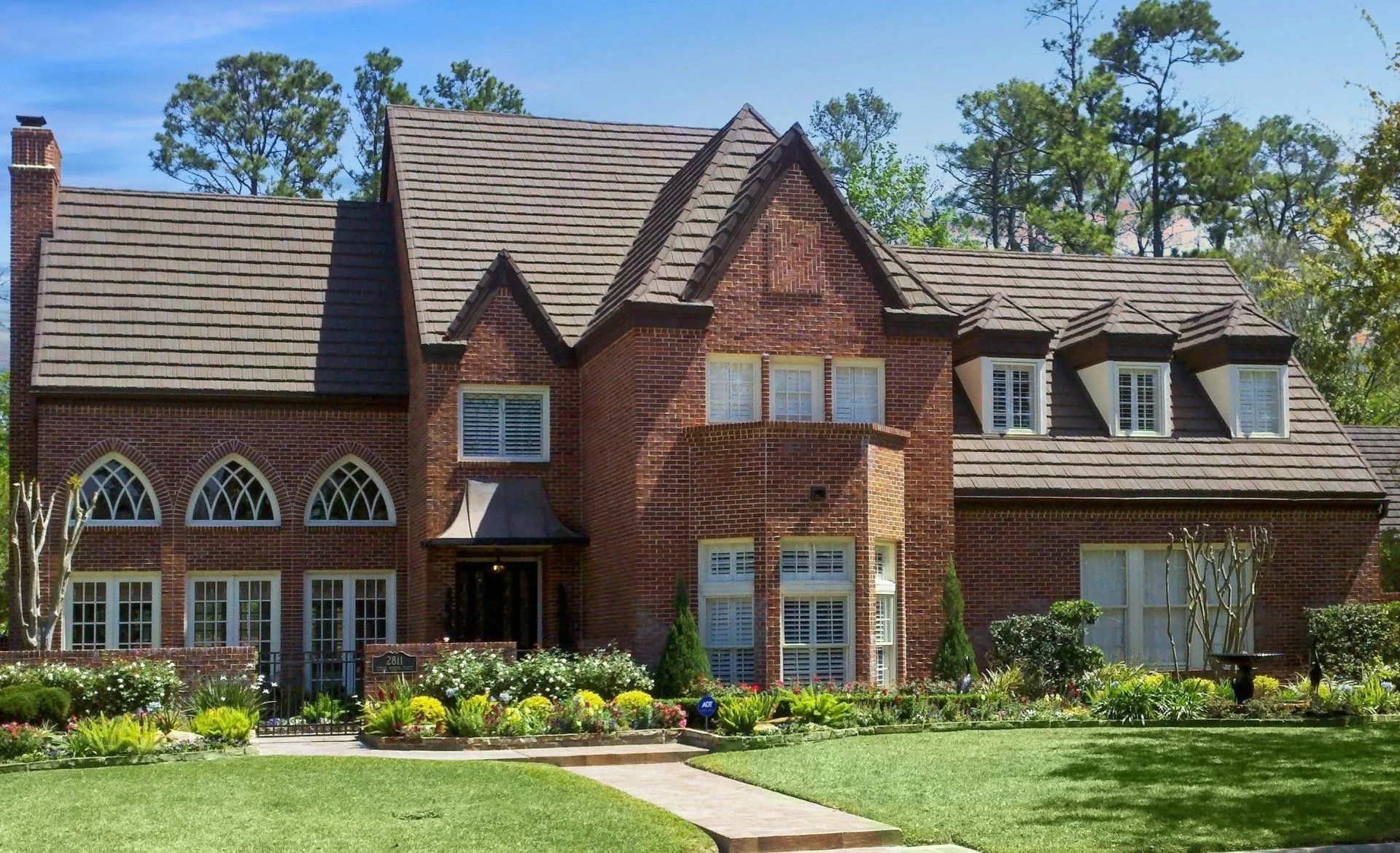 A two-story red brick house with a brown shingled roof, arched windows, and a landscaped front yard.