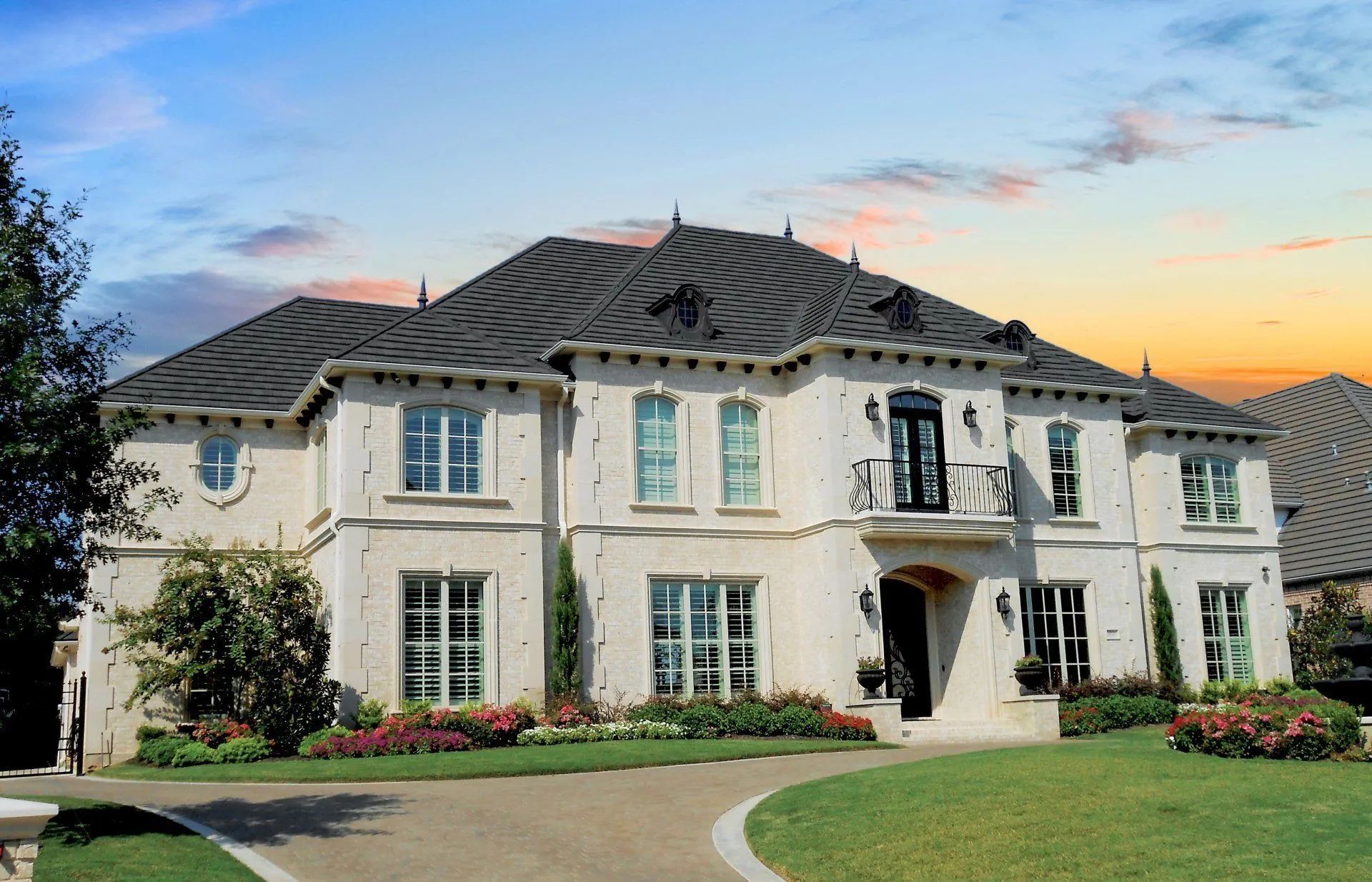A large, light-colored two-story luxury home with a dark, multi-gabled roof and a paved driveway at sunset.