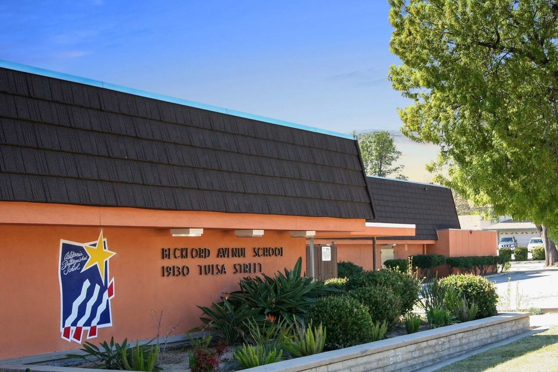 An exterior view of Beckford Avenue School with orange walls, a brown shingled roof, and a blue and yellow star emblem.