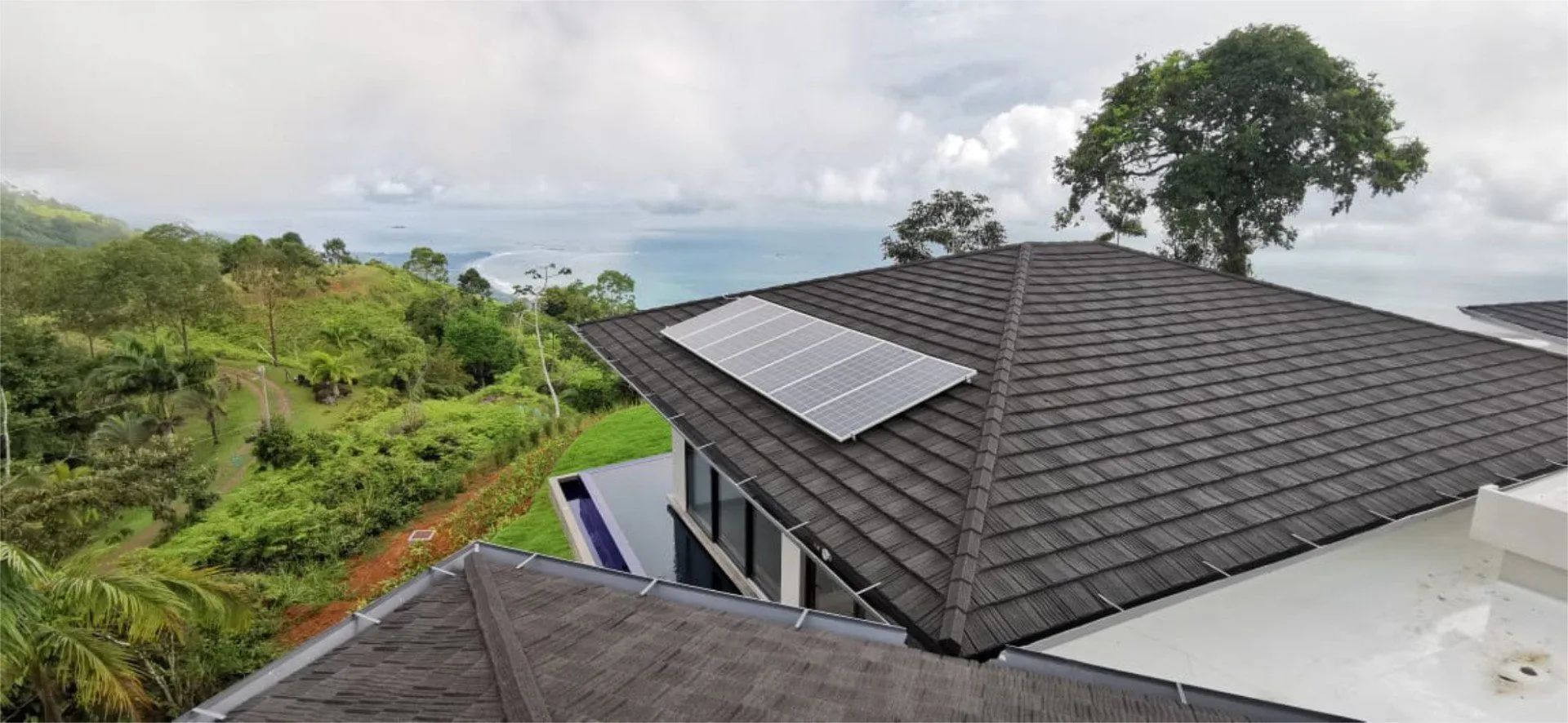 A high-angle view of a dark shingled roof with a rectangular solar panel array, overlooking a lush, hilly coastal landscape.