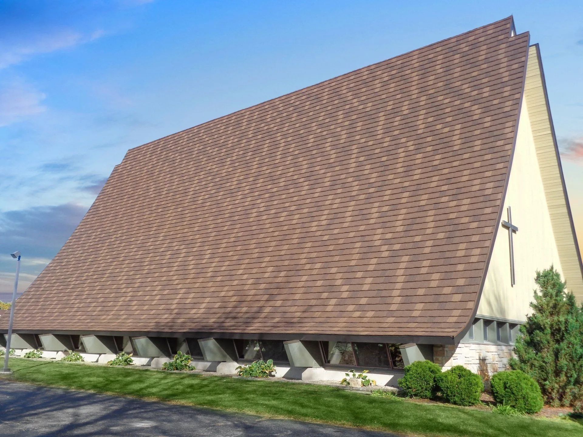 A-frame church with a large brown shingled roof, light-colored side walls, and a small cross near the front entrance.