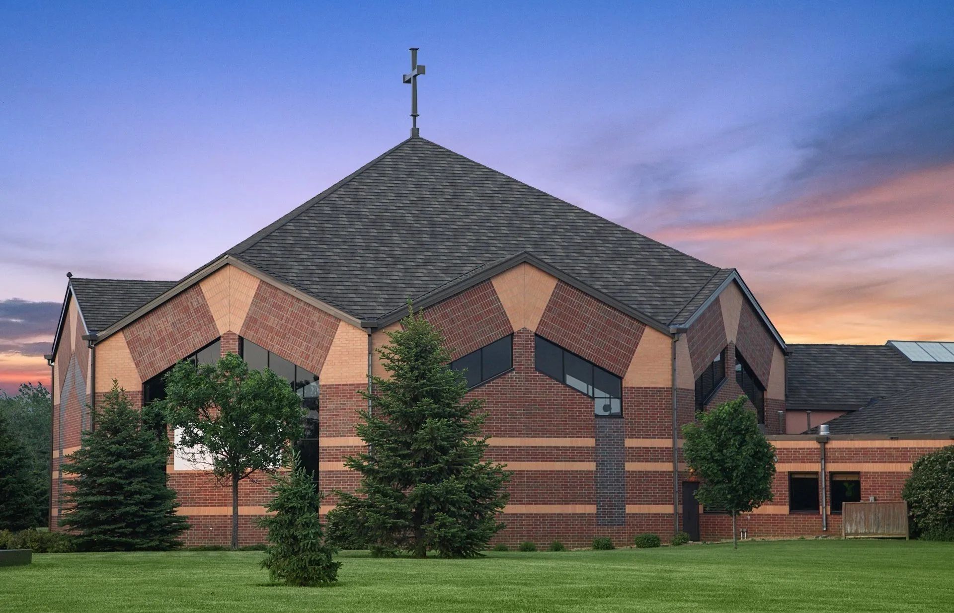 A modern brick church building with a cross on its roof, set against a sunset sky.