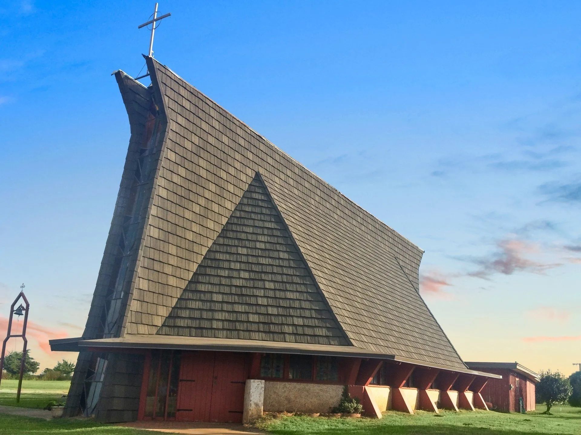 A modern church building with a large, sharply peaked, shingled roof and red doors, set against a blue, sunlit sky.