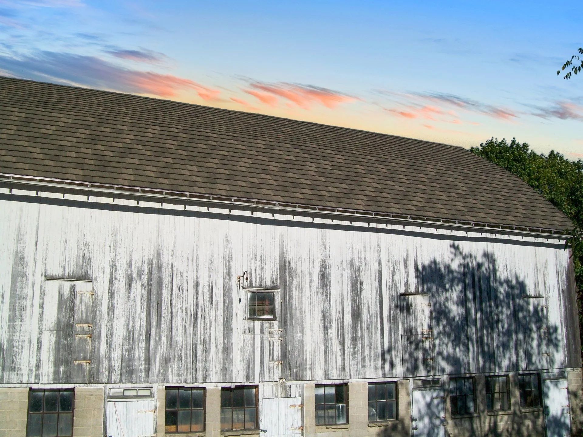 A white, weathered barn with a shingled roof under a colorful sunset sky.