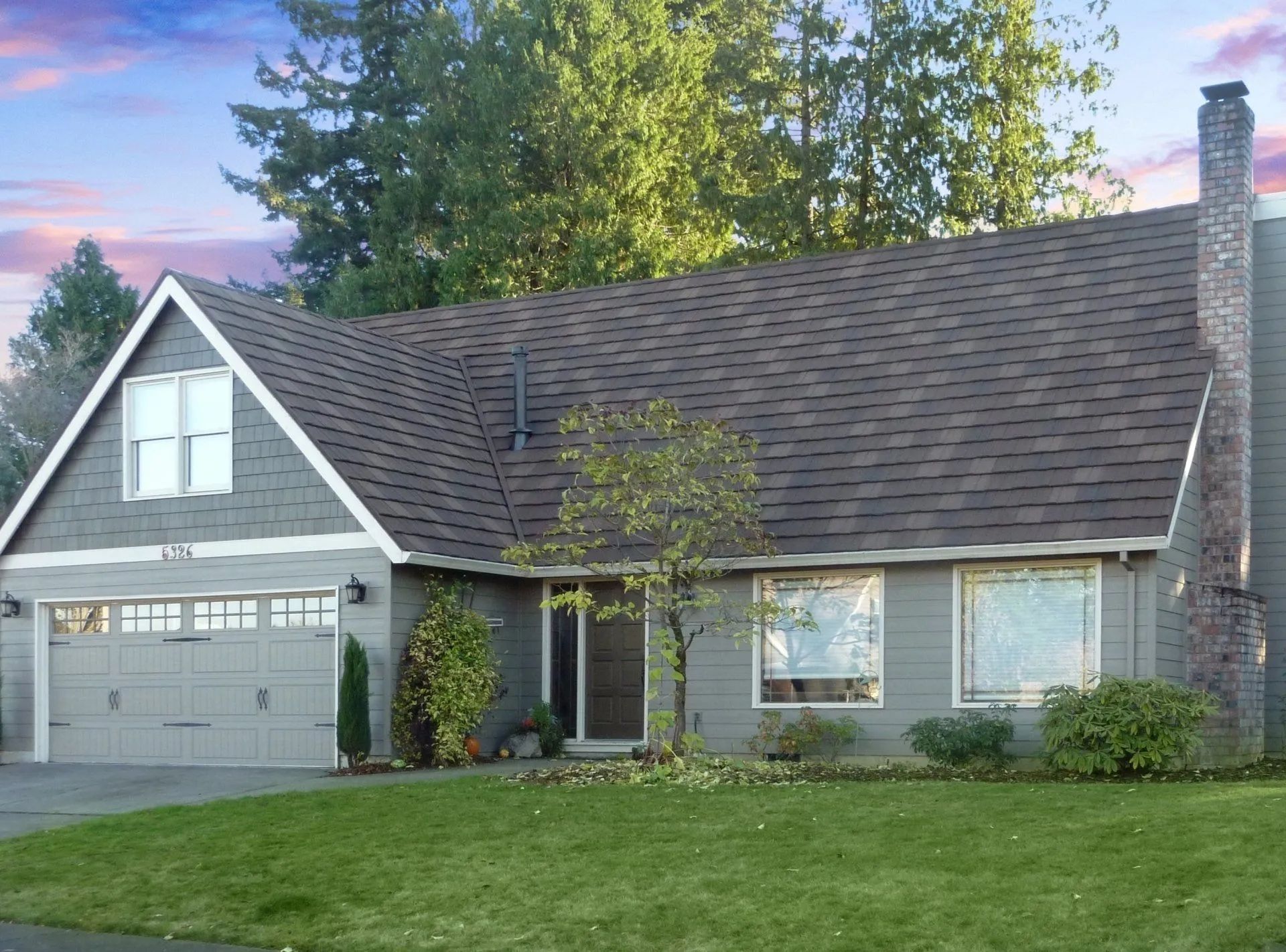 A two-story grey suburban house with a brown shingled roof, a front-facing garage, and a manicured green lawn.
