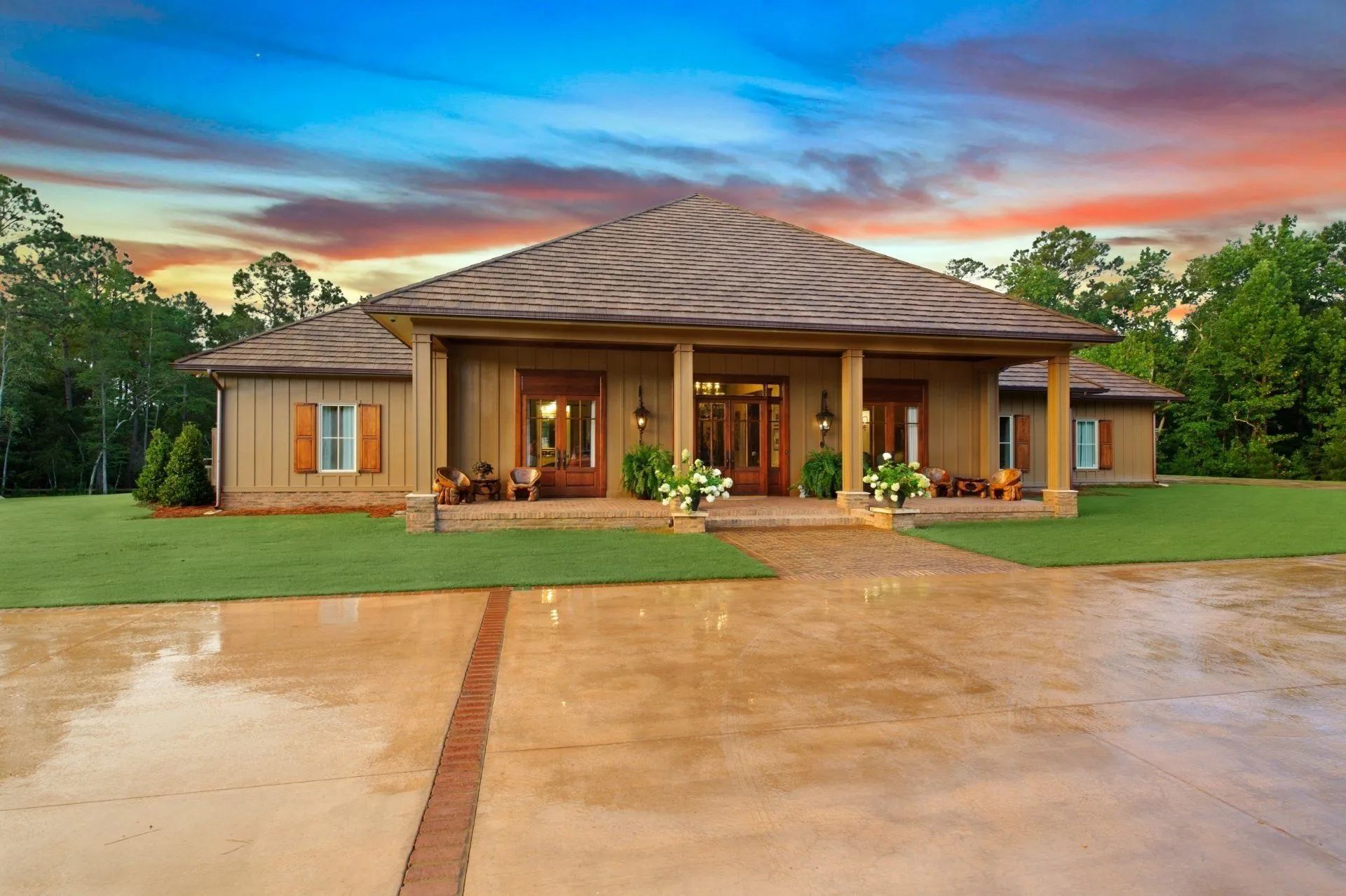 A tan, single-story house with a hip roof and columned porch sits behind a large, wet concrete driveway at sunset.