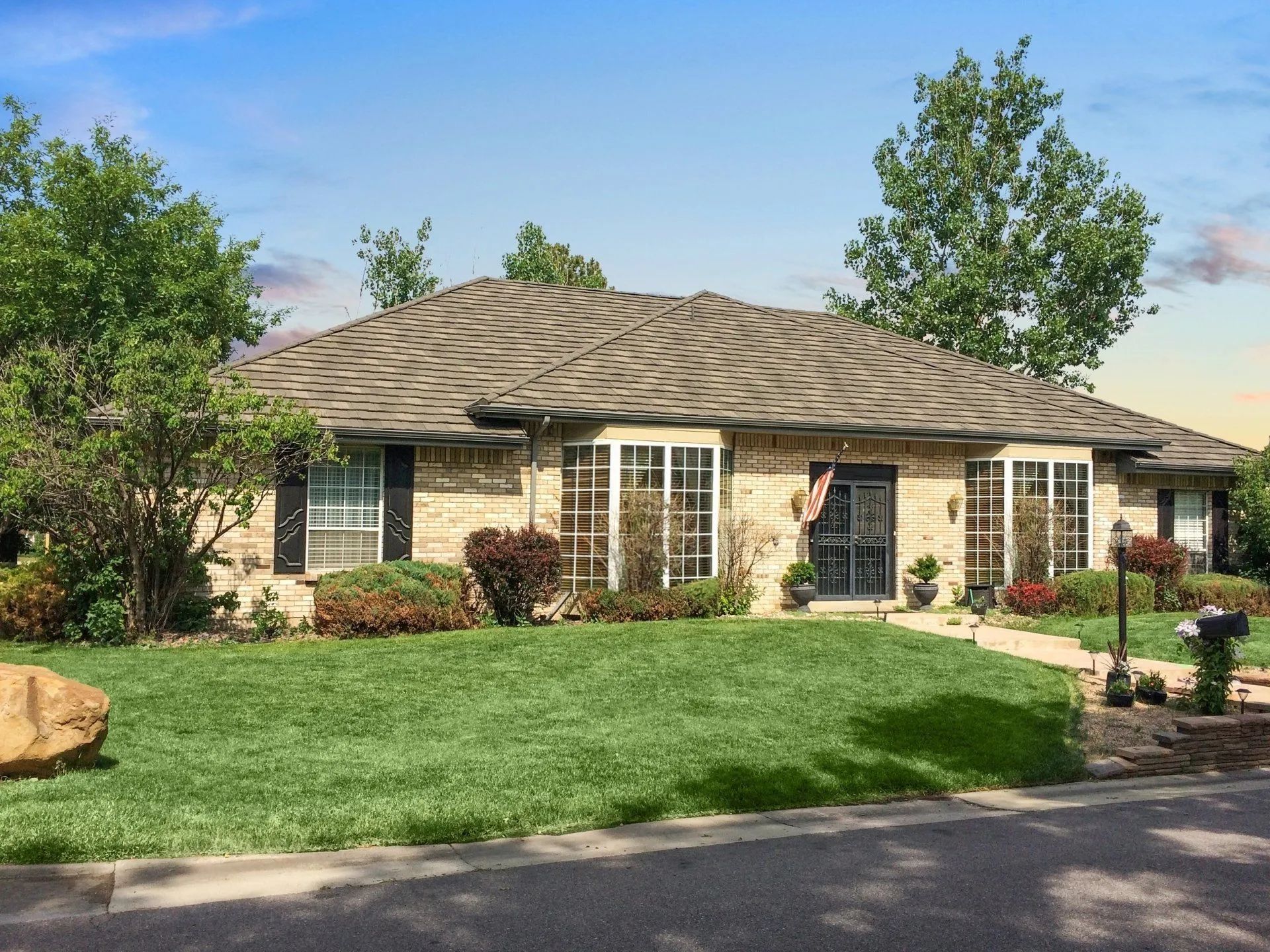 A single-story, beige brick house with a dark, patterned roof, lush green lawn, and trees under a clear blue sky.