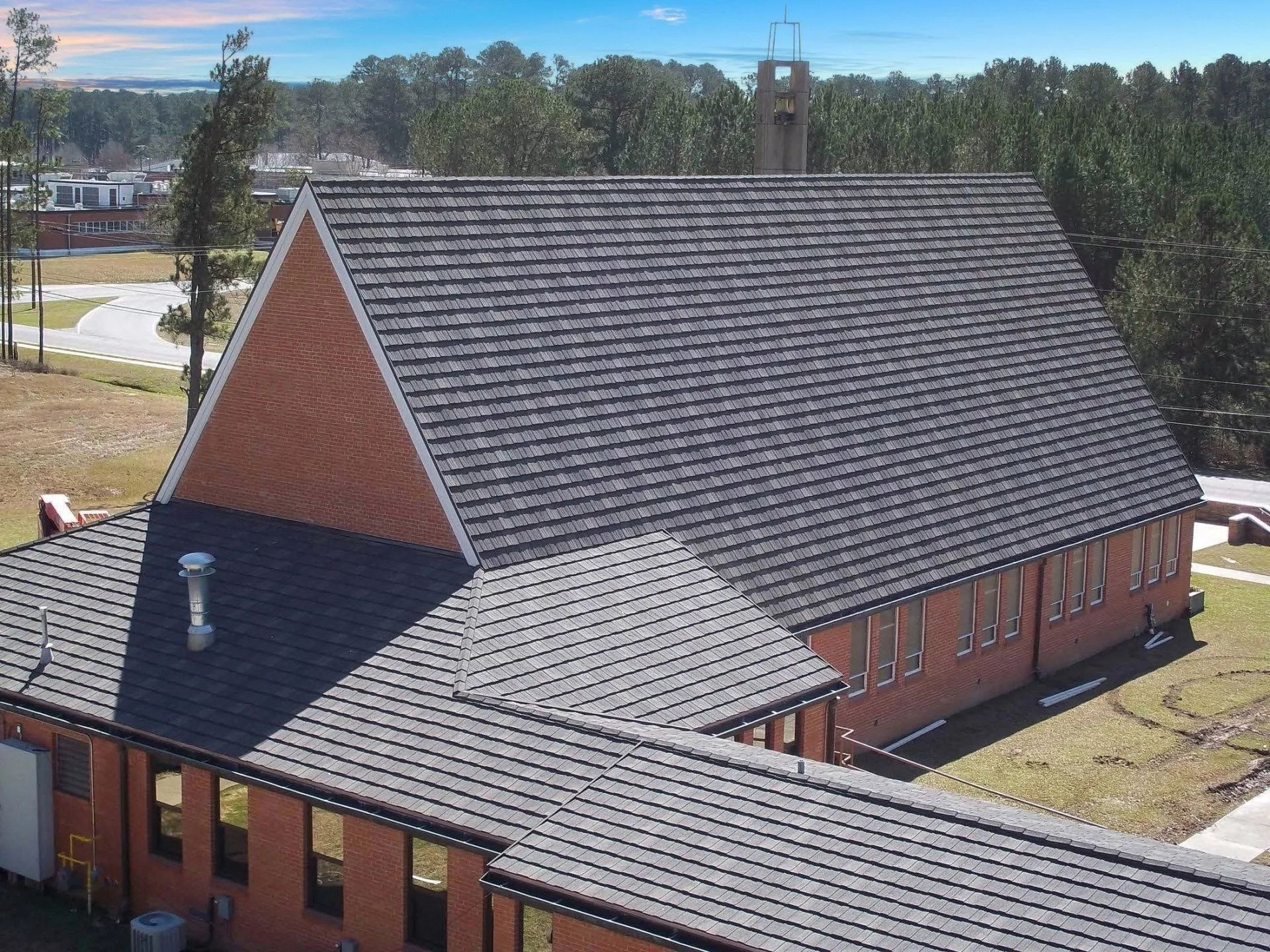 High-angle view of a brick building with a dark shingled roof, set against a backdrop of trees and a bright blue sky.