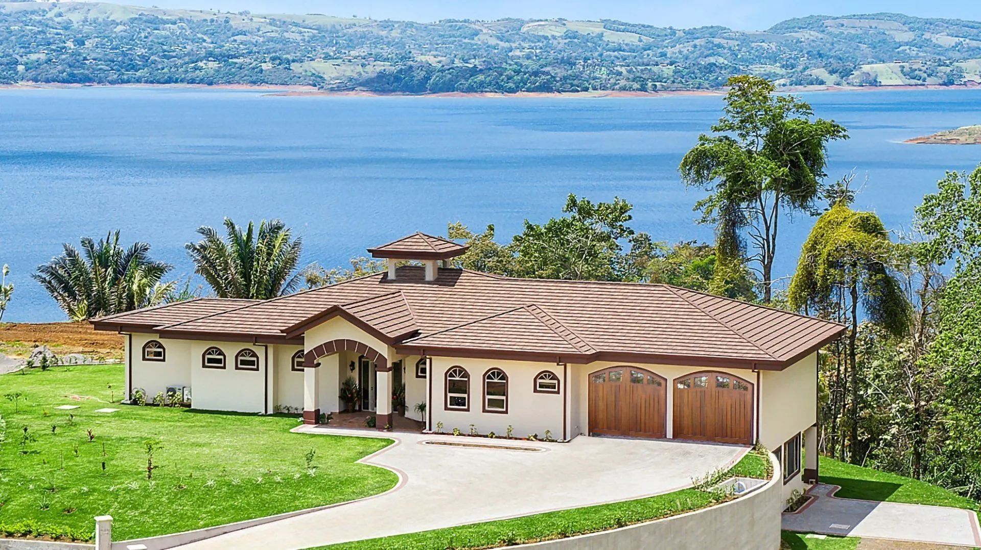 Single-story stucco house with a tiled roof and two-car garage, overlooking a large lake and distant hills.