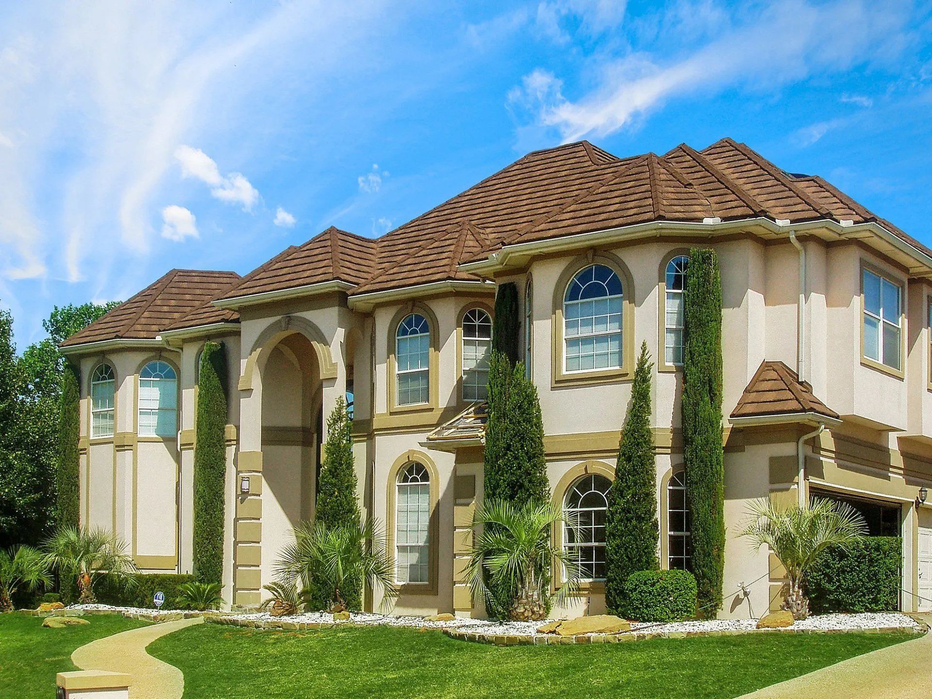 A large, multi-story beige stucco house with a brown tiled roof, arched windows, and tall evergreen trees under a blue sky.