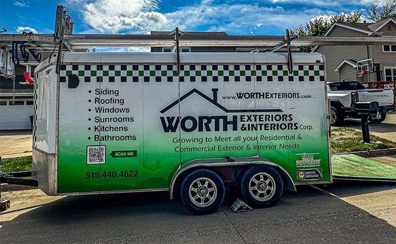 A white utility trailer parked on a residential street, branded with green logos for Worth Exteriors & Interiors.
