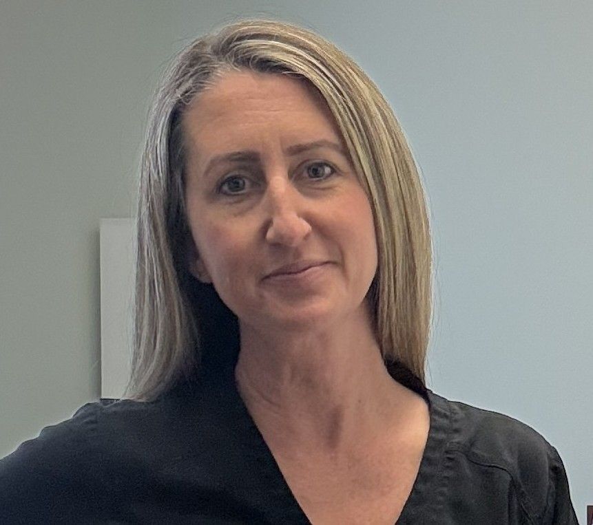 Woman in a black top standing against a plain gray wall, looking at the camera with a slight smile