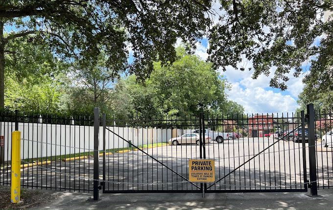A black gate with a yellow sign on it is surrounded by trees and a parking lot.