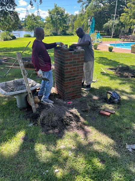 Two men are working on a brick chimney in a yard.