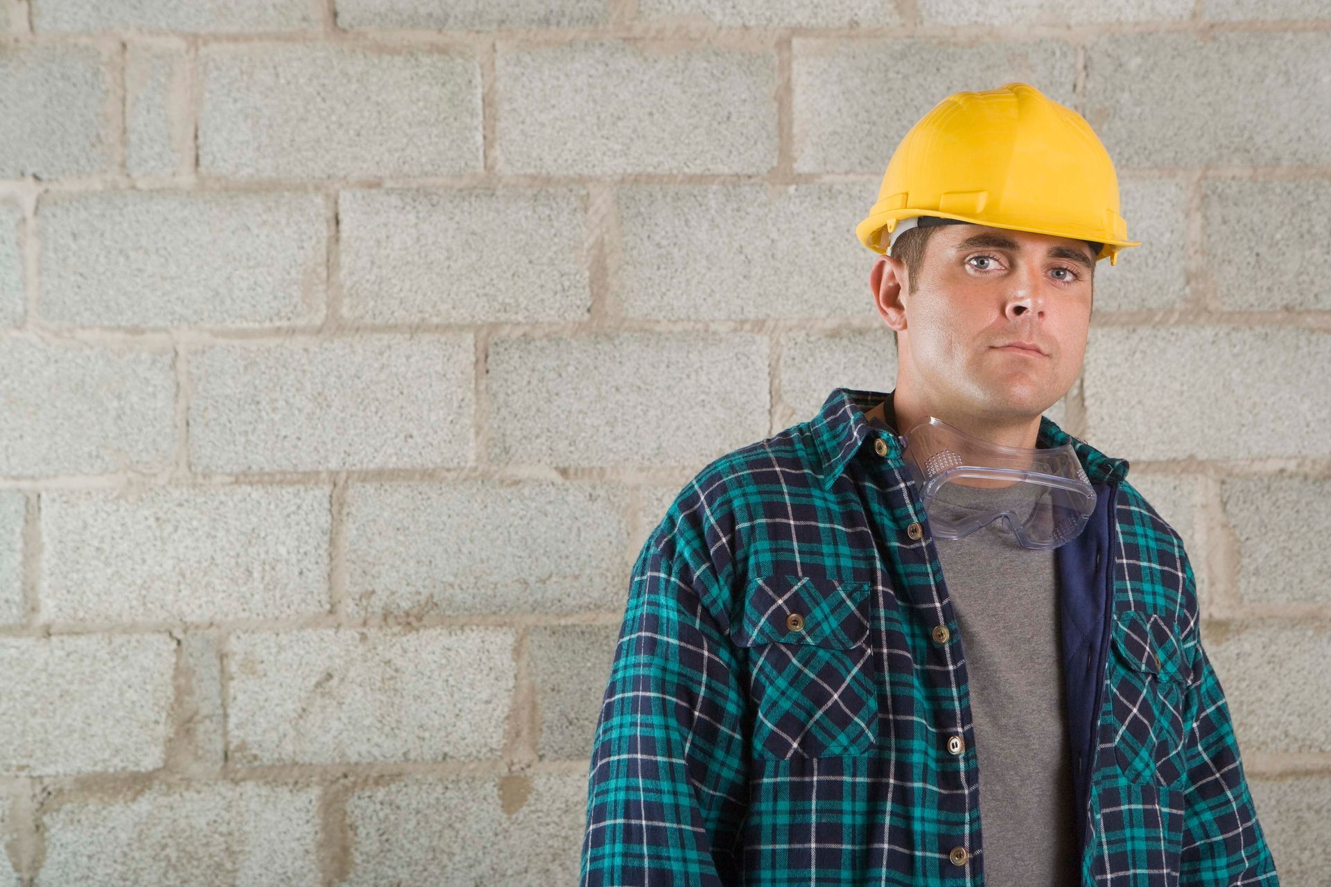 Man in yellow hard hat and plaid shirt stands in front of a concrete block wall.