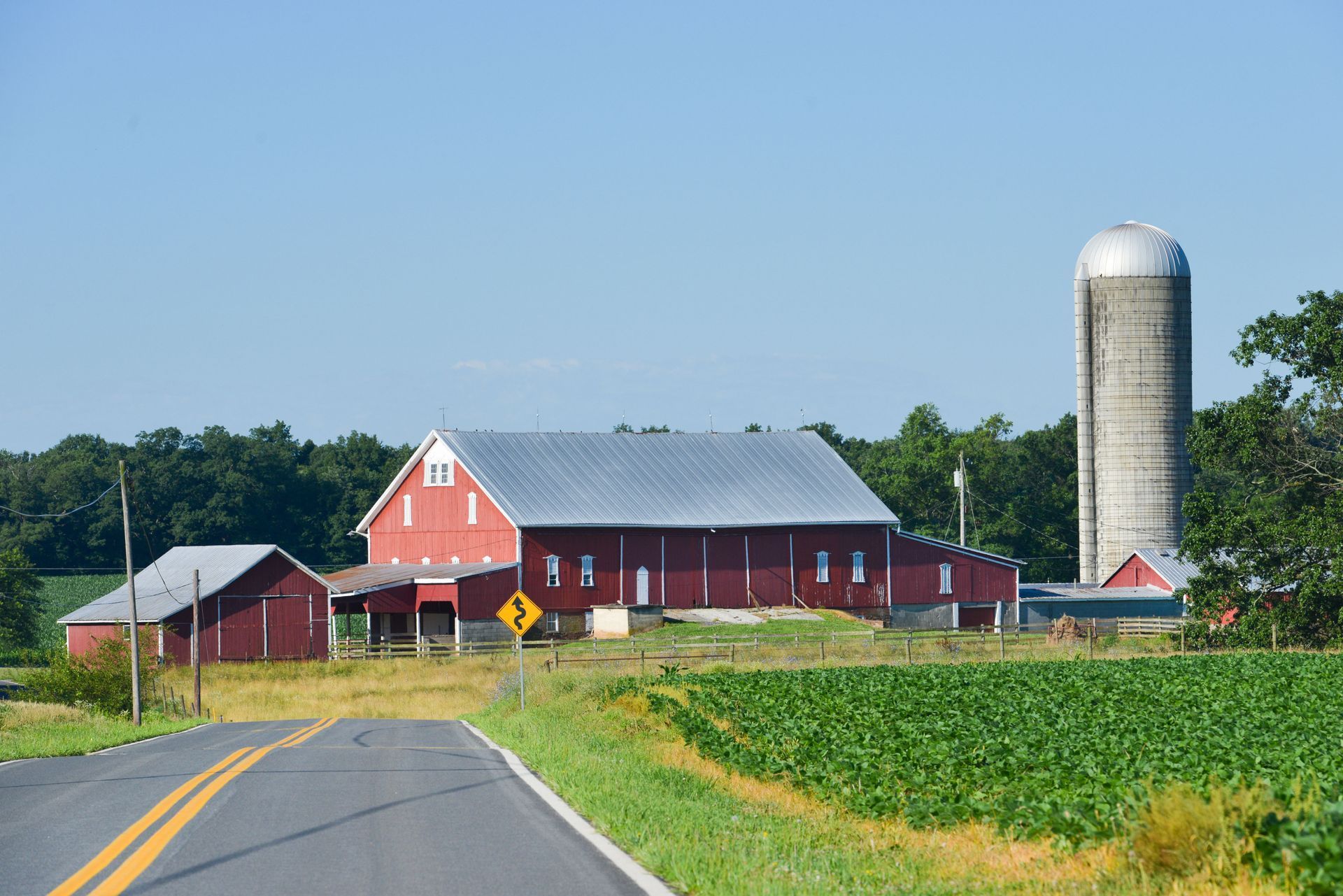 Red barn and silo on a sunny day next to a road and green fields.