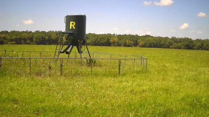 Black deer feeder on stilts in a fenced grassy field under a cloudy sky