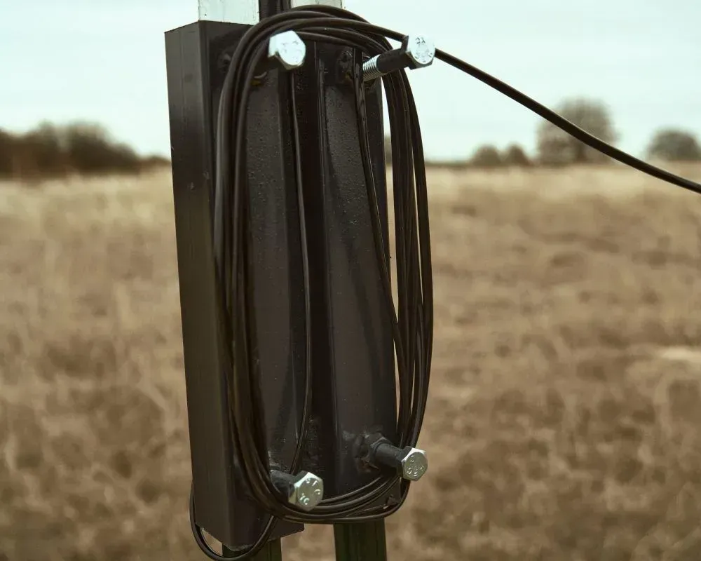 Black metal box on a fence post holding looped black wire, with a field in the background.