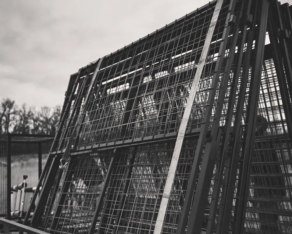 Metal cage structure, outdoors, angled perspective, grid pattern, against a blurred background of trees and sky.