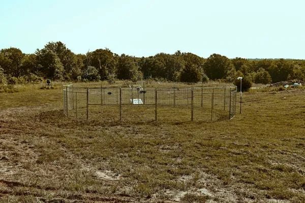 Circular fenced enclosure in a grassy field, trees in the background, clear sky above.