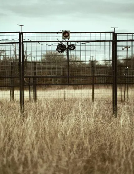 Black wire fence in tall, dry grass. Cloudy sky in the background. Warning sign attached to the fence.